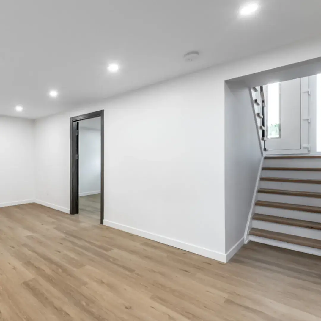 Newly renovated basement with white walls, wooden flooring,and a staircase leading to the main floor.