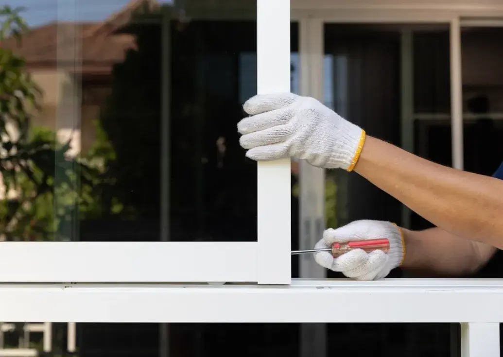 Worker wearing white gloves installing a white aluminum window frame using a screwdriver.