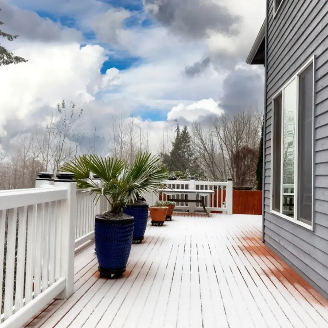 A wooden deck covered in light snow, featuring potted plants and a white railing.