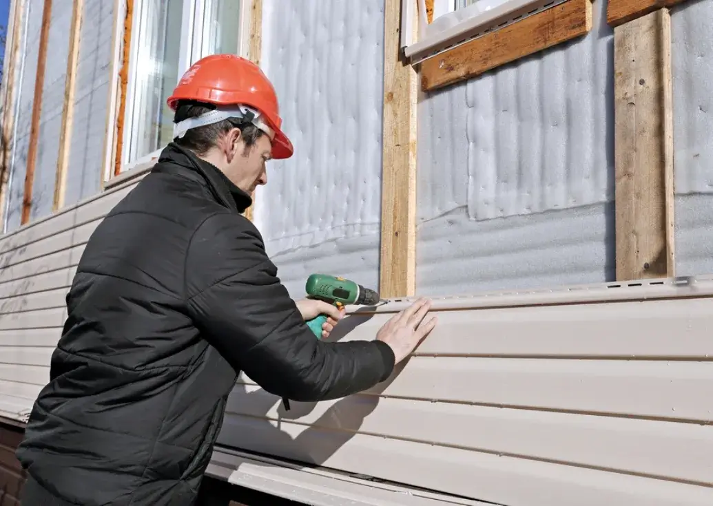 Construction worker in a red helmet installing vinyl siding on the exterior wall of a house using a power drill.